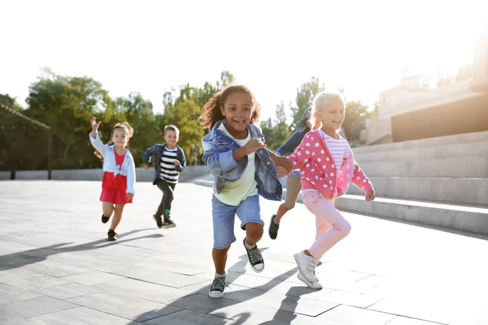 Group of five small children running on concrete outdoors on sunny day.